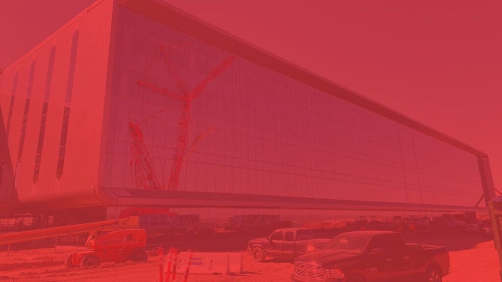 Large building under construction with a reflective glass facade, multiple cranes, and construction vehicles at the site. The image, captured by Tilden, is heavily tinted red.
