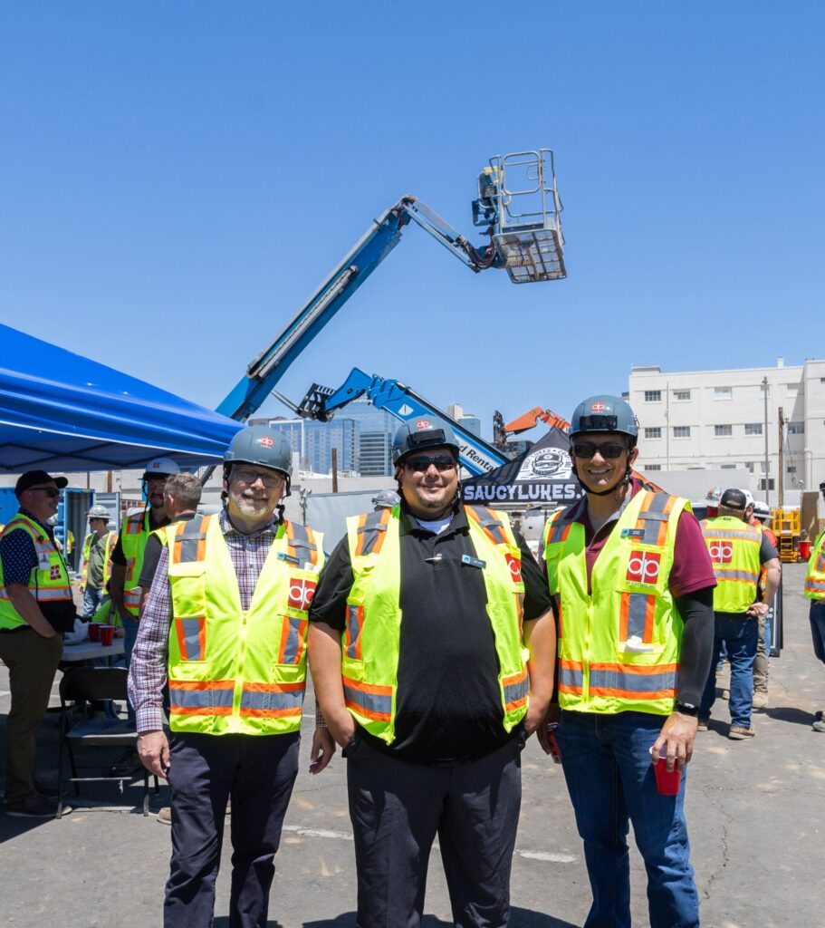 Three construction workers wearing hard hats and reflective vests stand in front of a blue canopy with lift equipment and other workers in the background on a sunny day. DP Electric is committed to safety and support, especially during Mental Health Month.