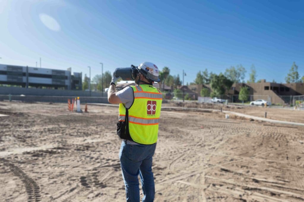 A construction worker in a neon safety vest and helmet, holding a surveying instrument on a tripod, stands at a large, cleared site with buildings and trees in the background. DP Electric ensures proper prevention measures are in place to combat heat illness on such demanding projects.