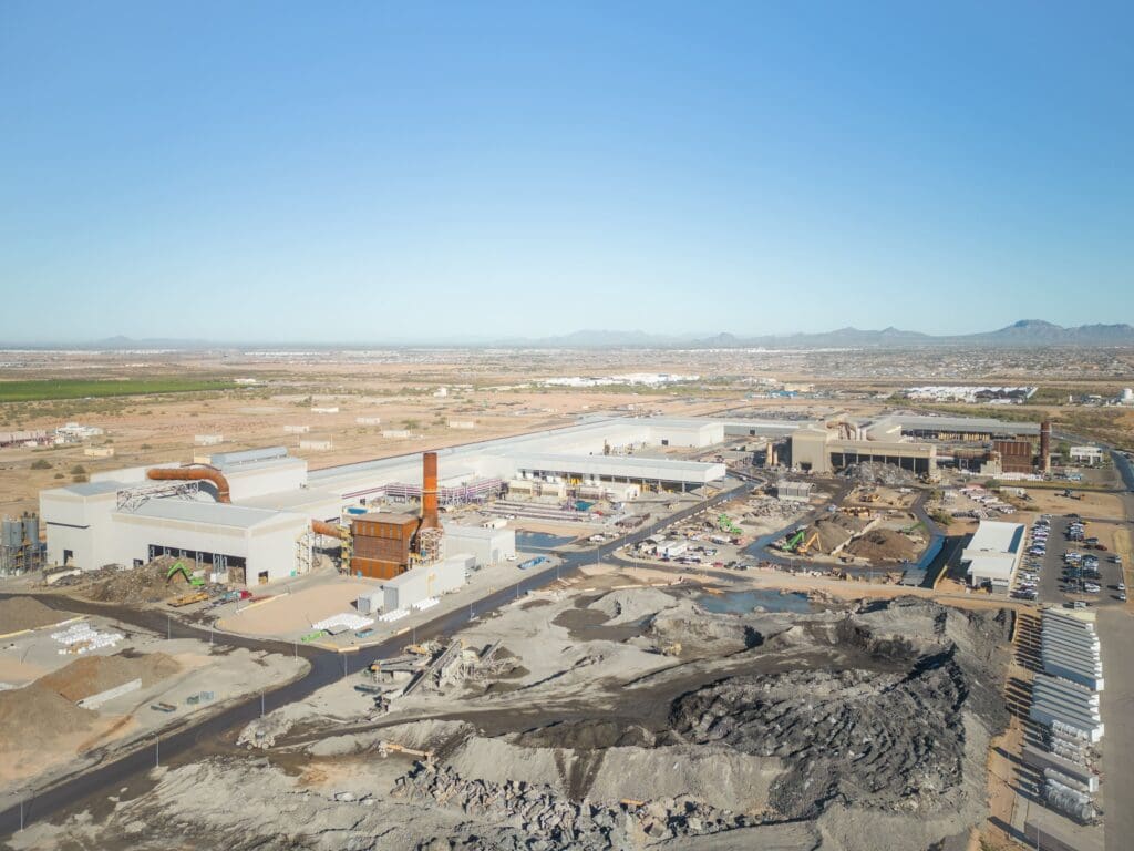 Aerial view of a CMC Steel industrial complex with multiple buildings and equipment, surrounded by barren land and construction sites under a clear blue sky.