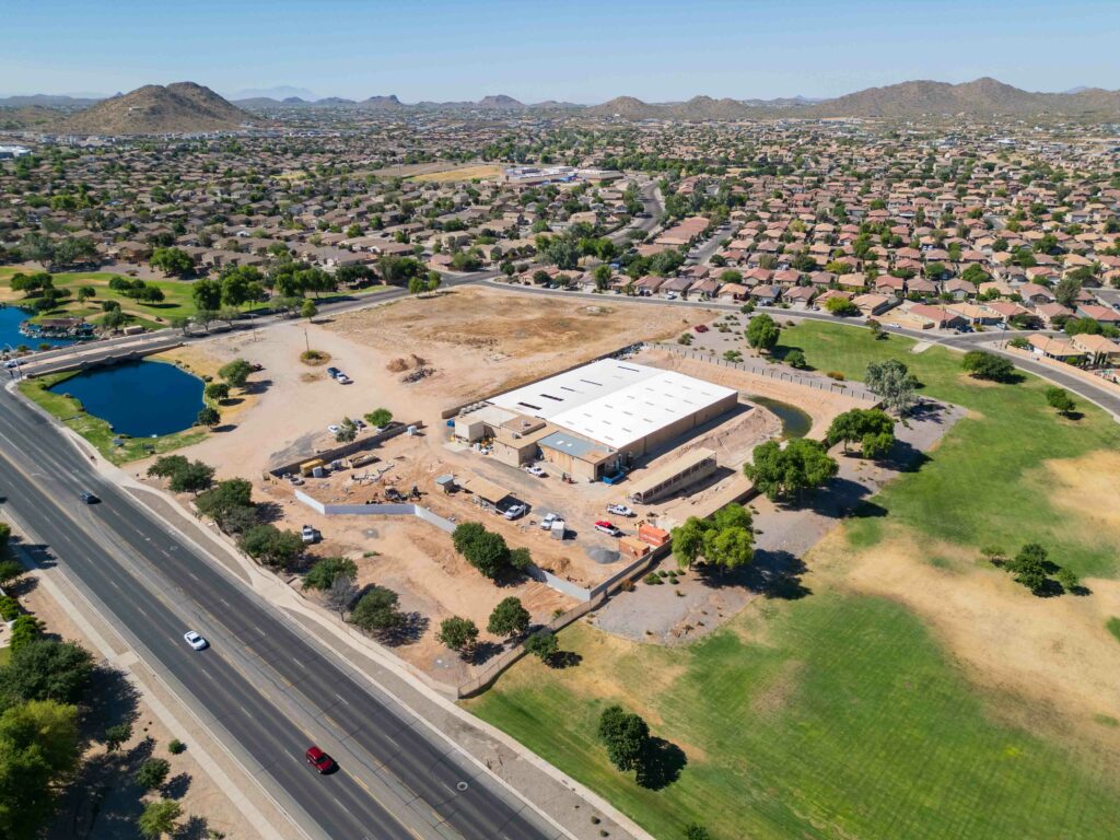 Aerial view of a suburban area featuring a large building under construction, surrounded by residential houses, a pond, a road, and green spaces.