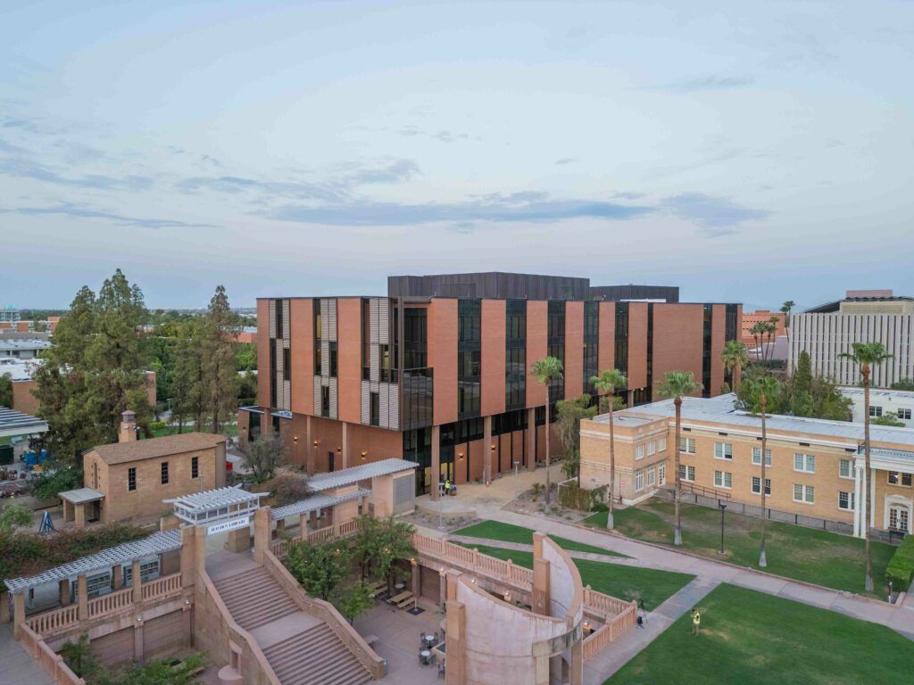 A contemporary multi-story building with horizontal windows, surrounded by trees and older buildings, is situated in a campus-like setting with pathways and green spaces.