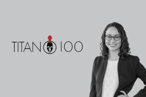 A woman in a business suit and glasses stands with her hand on her hip, smiling. Next to her is the 'Phoenix Titan 100' logo featuring a helmeted figure. The background is grey.