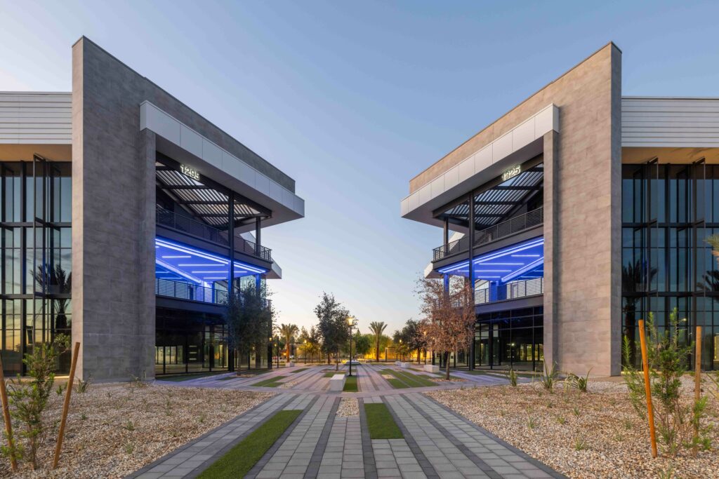 Modern architecture with two facing buildings featuring blue neon lights, separated by a landscaped pathway with trees and shrubs, set against a clear sky at dusk.