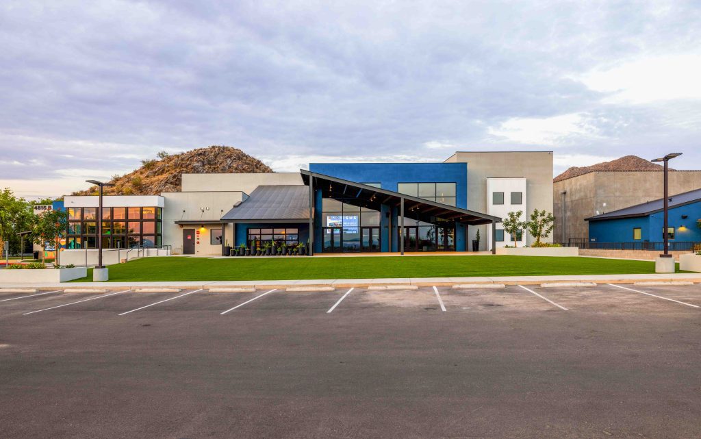 Modern commercial building with large windows, an angled roof, and a covered outdoor seating area, set in front of an empty parking lot and hills—perfect for a community space like Mountain View Church.