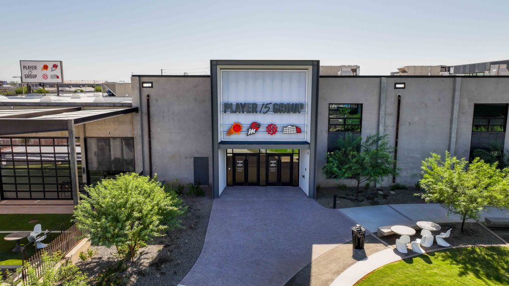 Front entrance of a modern building labeled "Player 15 Group" with glass doors, signage, surrounding trees, and outdoor seating.