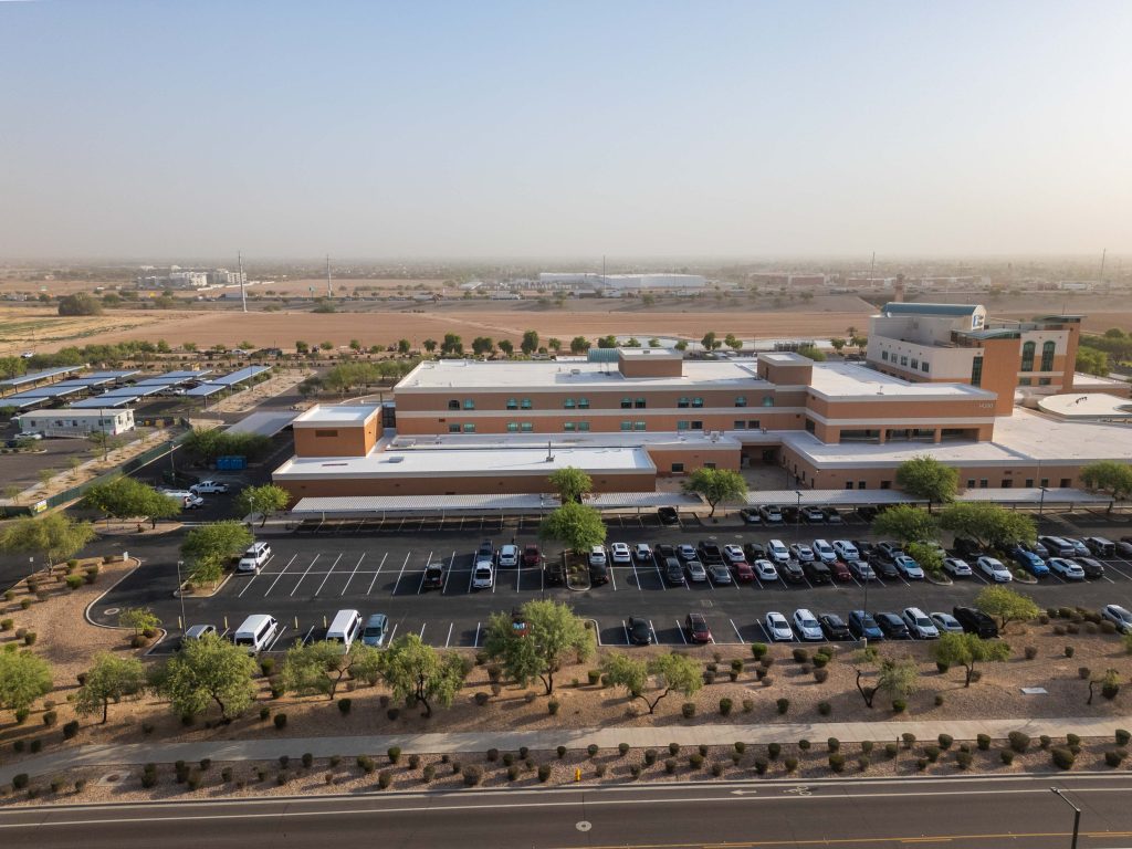 Aerial view of a large, modern Cancer Research Center building with a parking lot in front, surrounded by desert landscaping and sparse trees, under a clear sky.